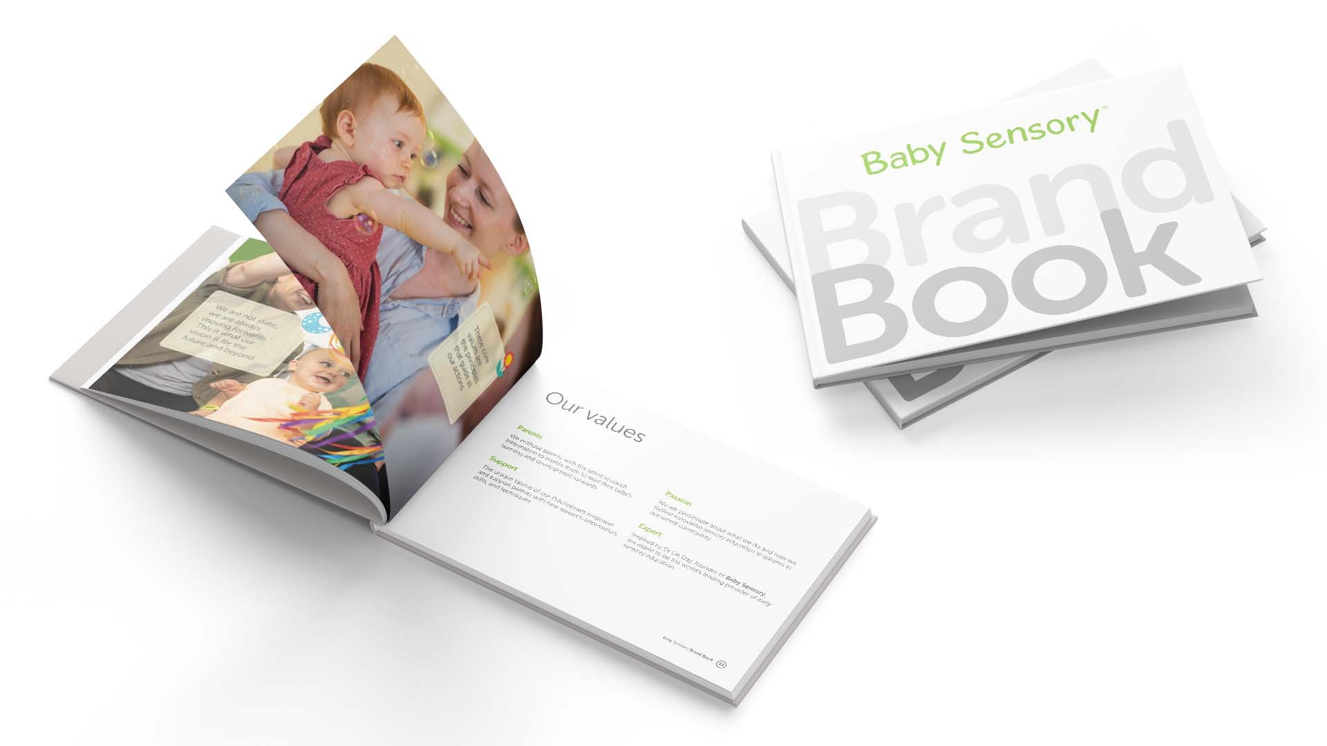 Open baby sensory brand book showing pages with images of a woman and young girl, and text about values like support, passion, and expertise, with additional stacked copies of the book on a white surface.