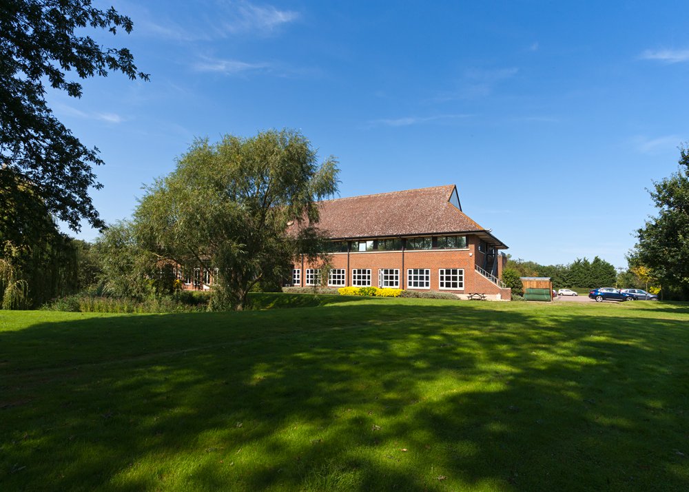 A brick building with a high-pitched roof, surrounded by green grass and trees, under a clear blue sky.