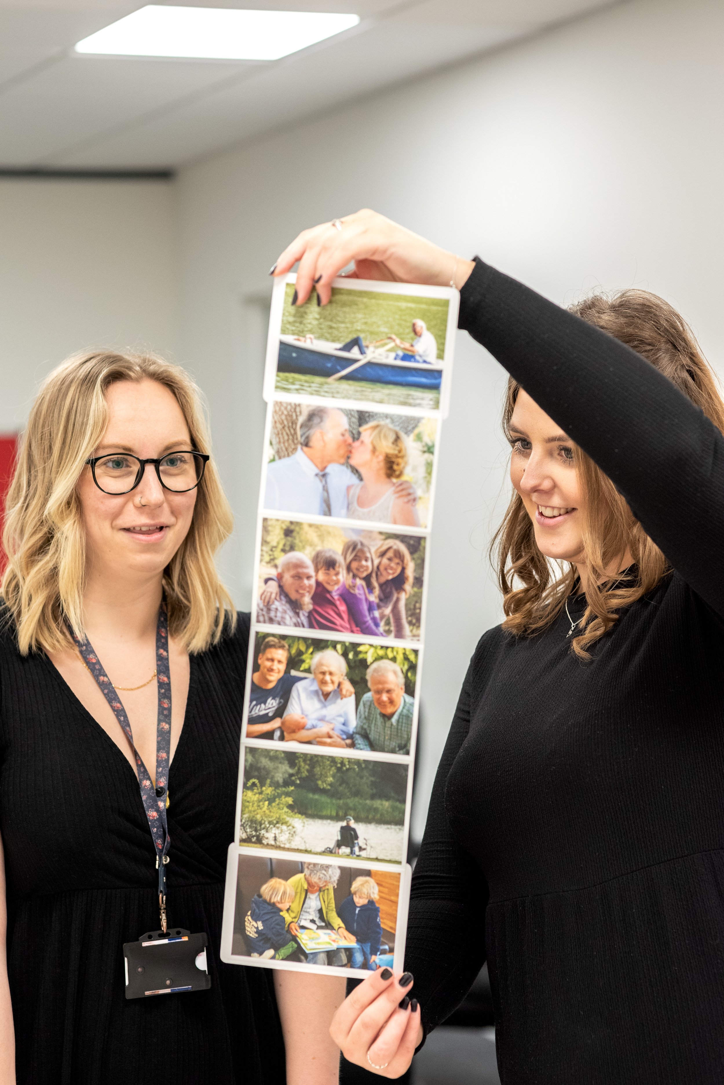 Two women examining a vertical photo collage featuring outdoor and family scenes in an indoor setting with plain walls and ceiling lights.