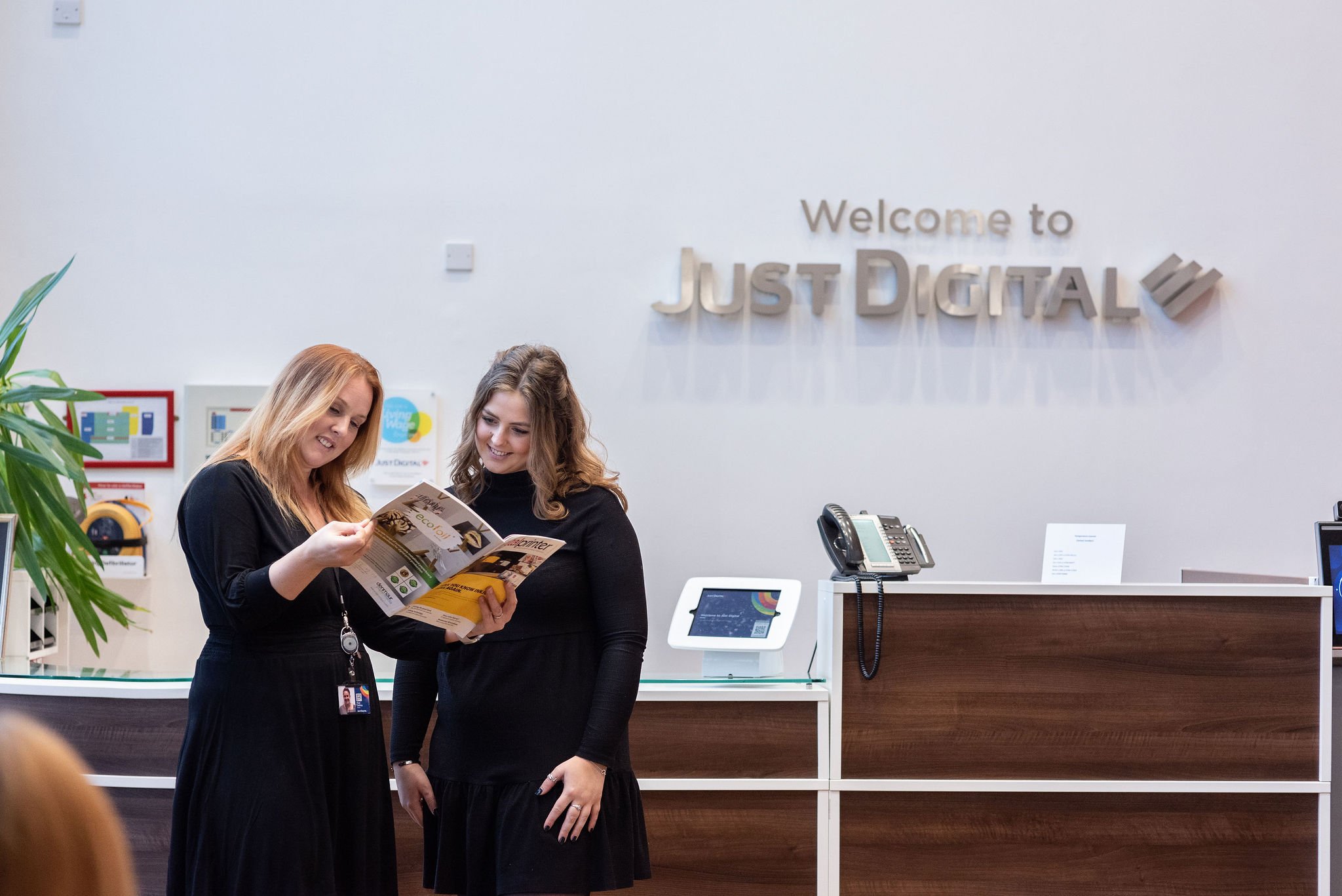 Two women in black dresses standing at a reception desk, looking at a brochure together, inside an office with a sign that says 'Welcome to Just Digital' on the wall.