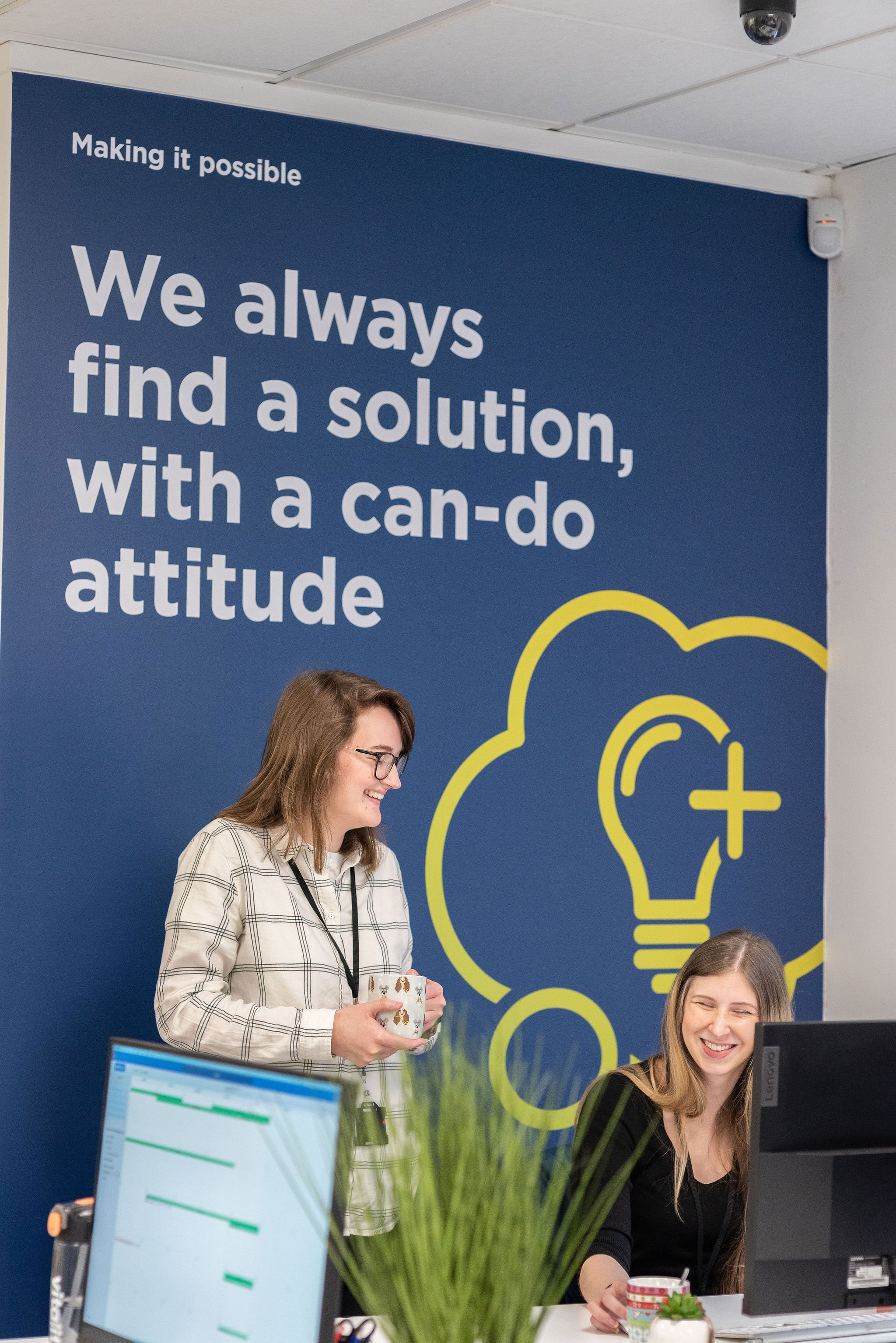 Two women in an office, one standing holding a mug and smiling, the other sitting at a desk looking at a computer screen and smiling. Behind them, a large blue wall has a motivational quote and a lightbulb graphic.
