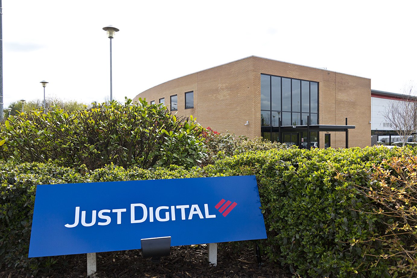 Exterior view of a modern building with large glass windows, surrounded by bushes, with a blue sign that reads 'Just Digital' in foreground.