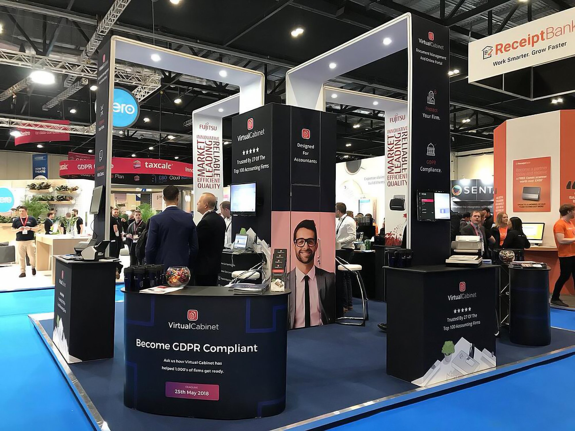 Trade show booth for Virtual Cabinet, a company specializing in GDPR compliance and accounting solutions, with banners, computers, and a few people engaging at the booth.