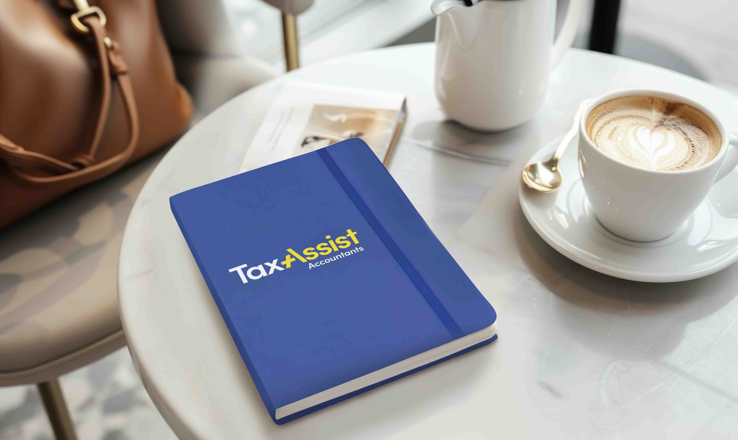 A white table with a blue accounting notebook labeled 'Tax Assist Accountants,' a cup of latte with foam art, a white teapot, and an open magazine. There is also a brown handbag nearby.