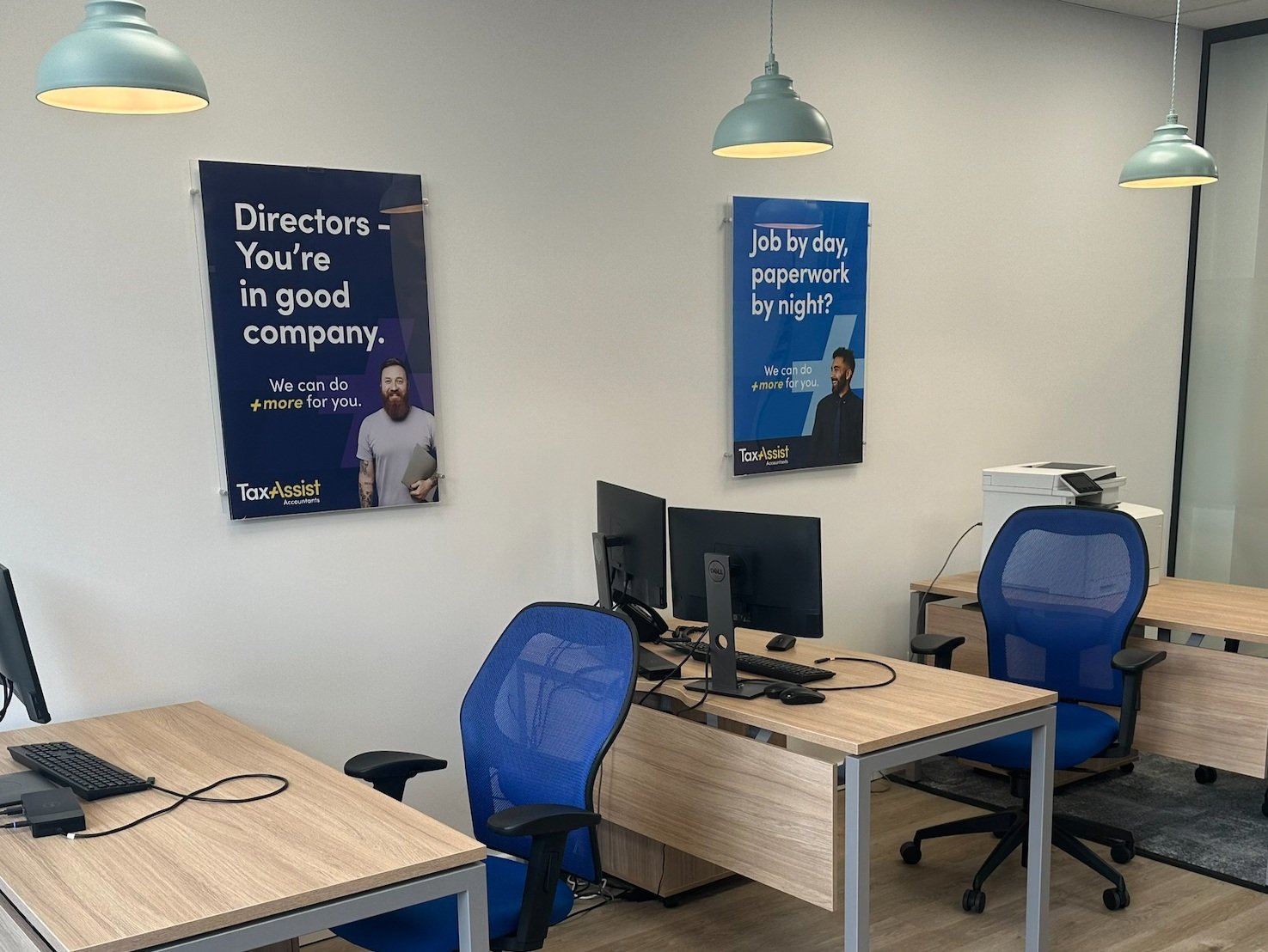 An office space with two desks, each with a computer monitor, keyboard, and mouse. There are two blue office chairs, one at each desk. On the wall, there are two posters promoting tax assistance services. The room has ceiling lights and a glass wall or door on the right side.