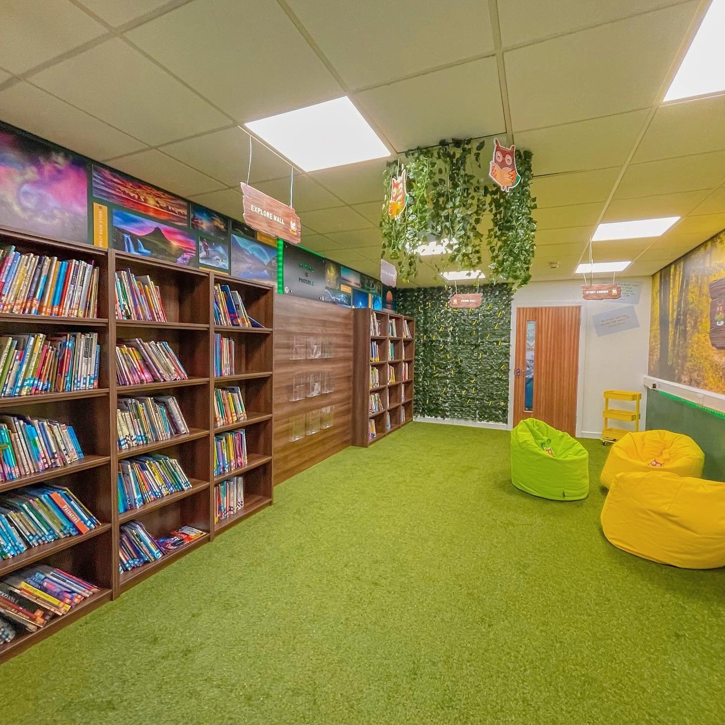 A children's library room with bookshelves, green carpet, green and yellow bean bag chairs, and decor such as hanging owl and fox cutouts, green vines, and wall art.