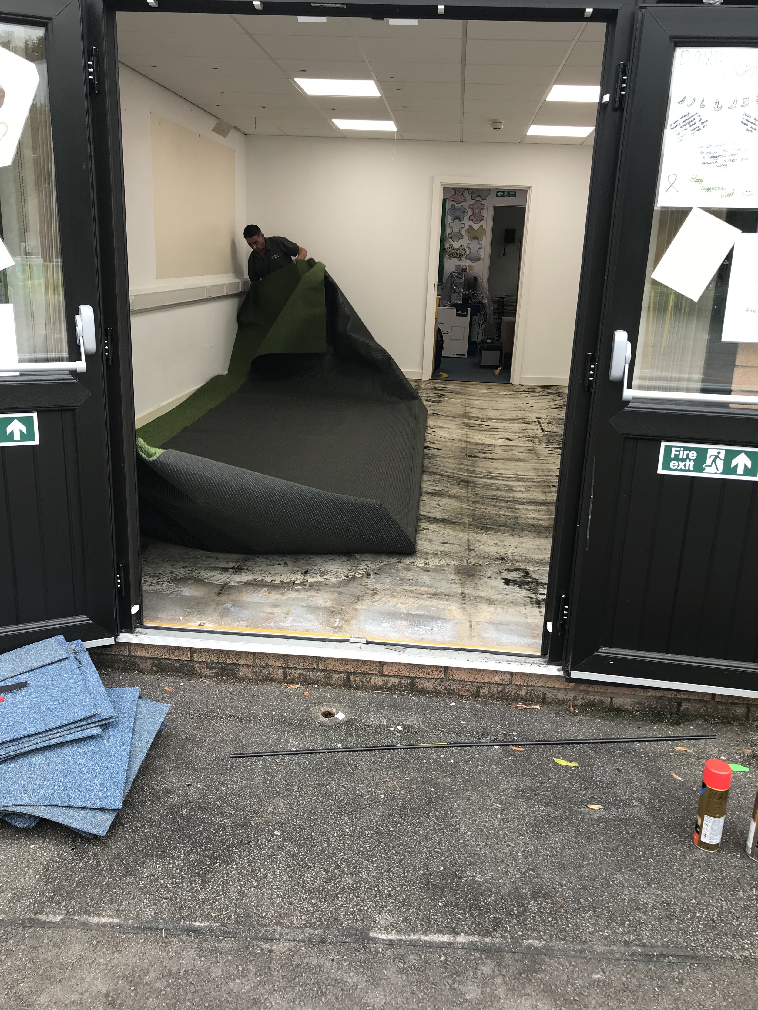 A person installing or removing a green and black carpet inside a room with a doorway and ceiling lights. The door is open, revealing the outside pavement and some flooring materials.