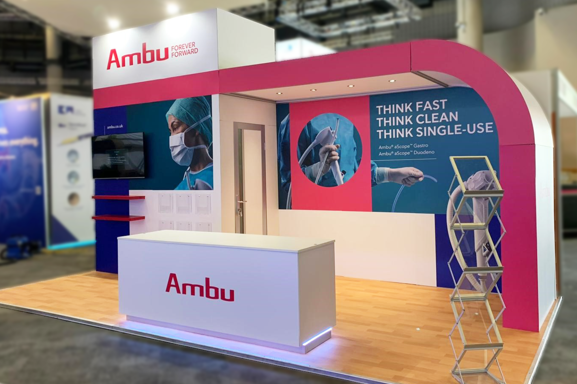 Trade show booth for Ambu with a pink and blue color scheme, featuring a desk with the Ambu logo, promotional posters of medical devices and a healthcare professional wearing a mask, and a foldable display stand in the front right corner.