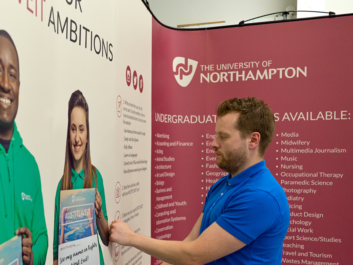 A man with a beard in a blue shirt pointing at a university poster. The poster features two smiling students and lists undergraduate programs at the University of Northampton.