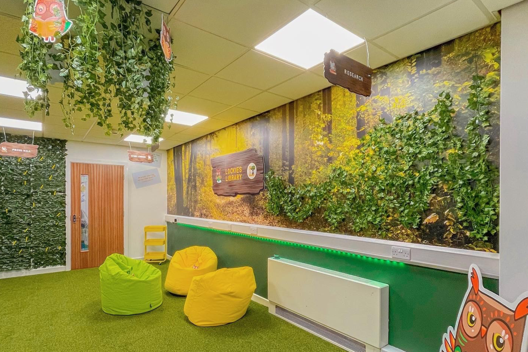 Children's library room with forest-themed wall mural, plush green and yellow bean bag chairs, artificial hanging vines, and signs labeled "Research" and "Lockies Library."
